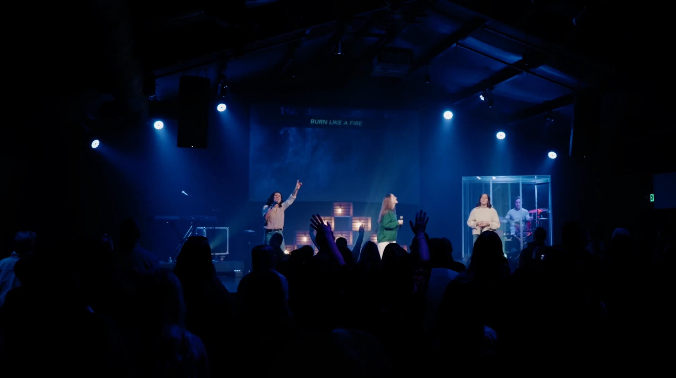 A band leads worship on a blue-lit stage. In the foreground, the silhouetted congregation raises their hands as lyrics display on a screen behind the band.